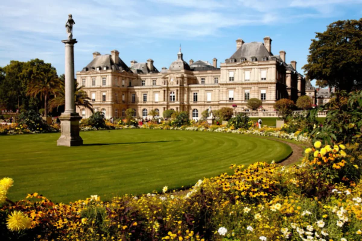 The Luxembourg Palace in Paris behind a curved lawn, a statue on a column and beds of yellow and white flowers