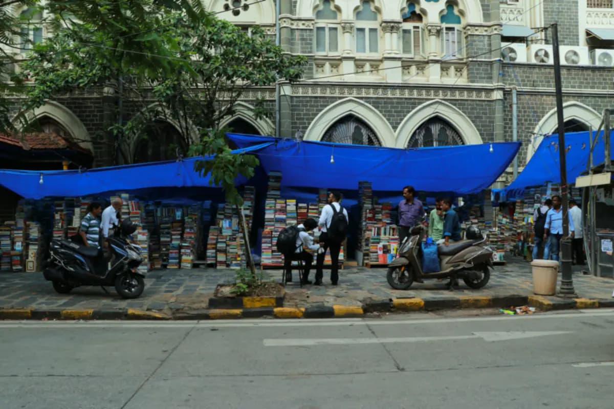 Stacks of books under blue tarpaulins on a Mumbai pavement, with people browsing in front of a Gothic style stone building