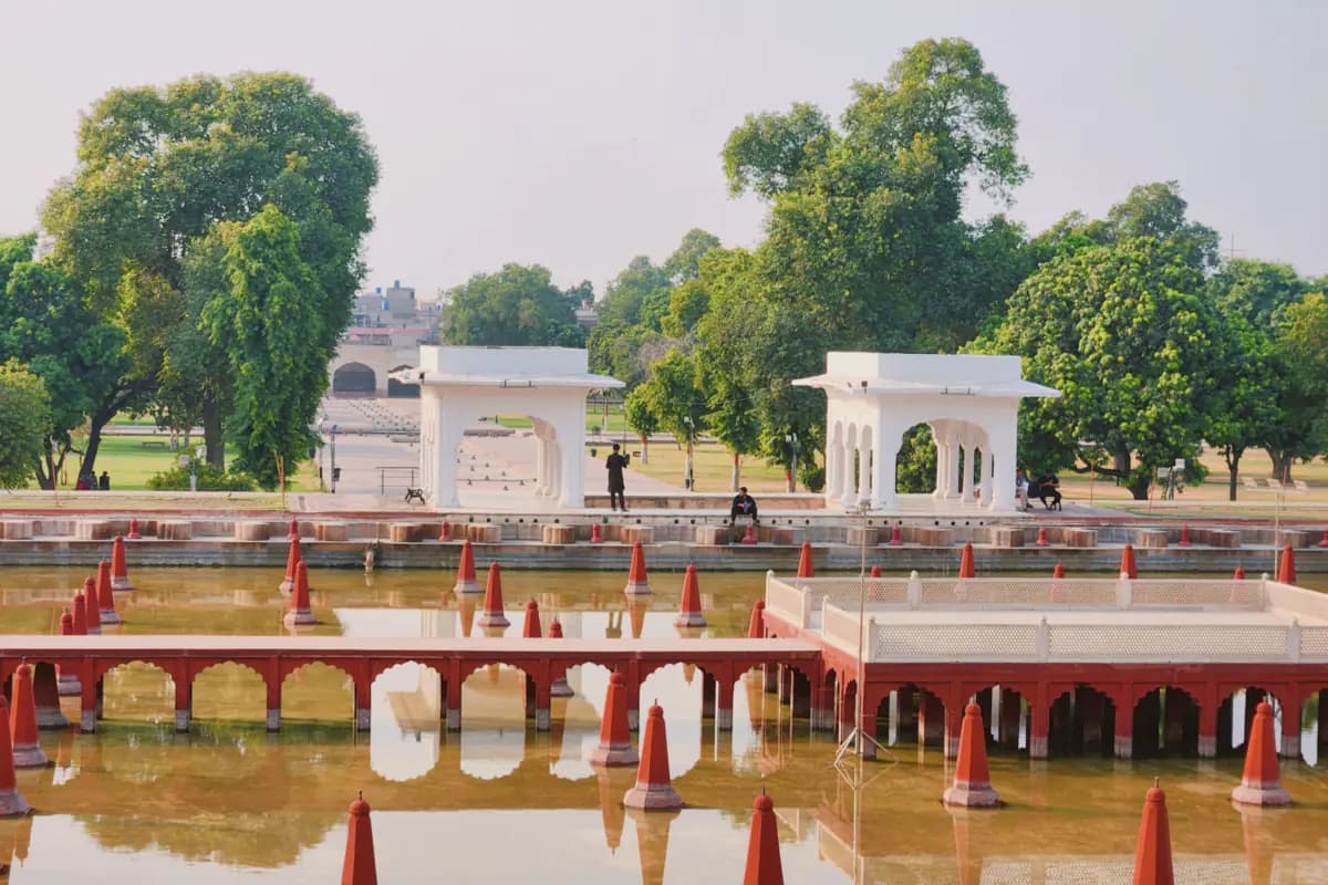 A water tank lined with red fountain spouts, white marble pavilions and tall trees in the Shalamar Gardens, Lahore