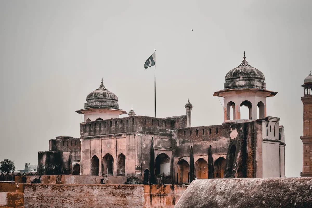 Domed pavilions and weathered walls of the Lahore Fort under a pale sky, with Pakistan's flag flying above
