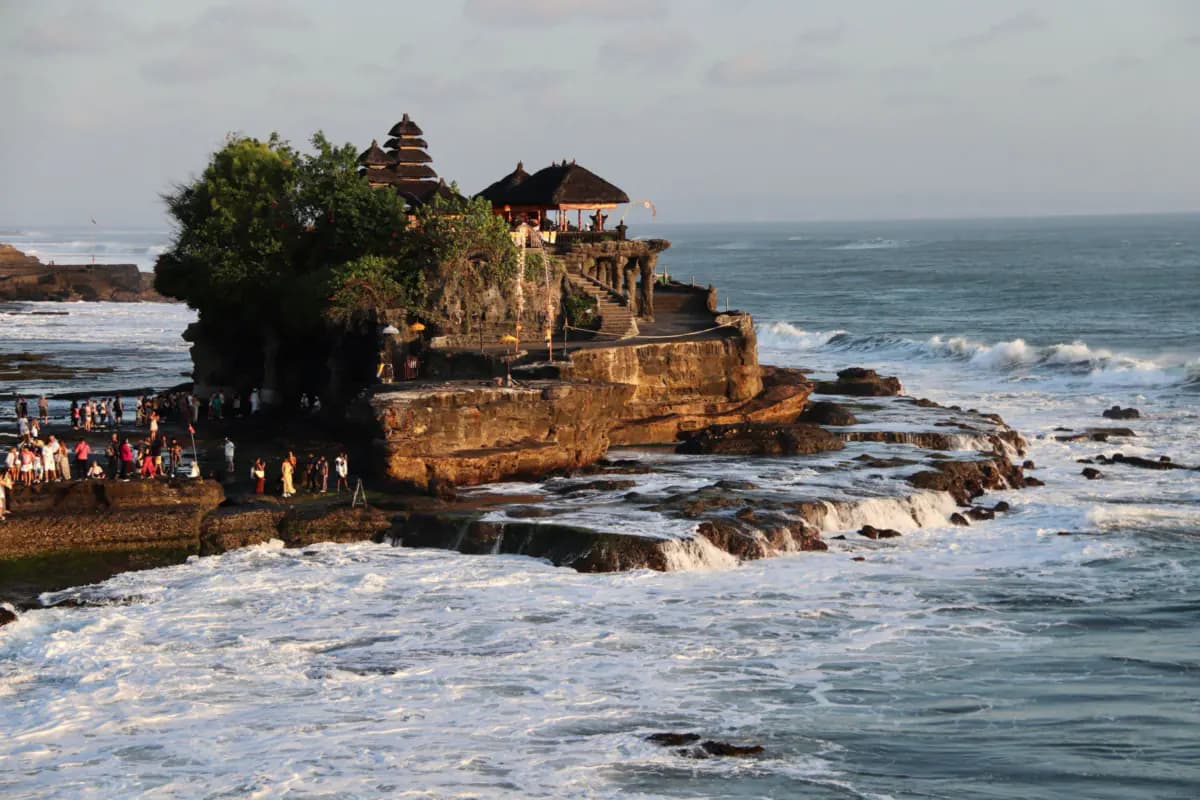 Tanah Lot temple with tiered shrine roofs on a rock outcrop, waves breaking around it and visitors on the shore