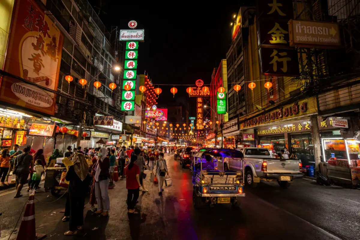 A street in Bangkok's Chinatown at night, with neon signs, red lanterns, people walking and a tuk tuk on the road
