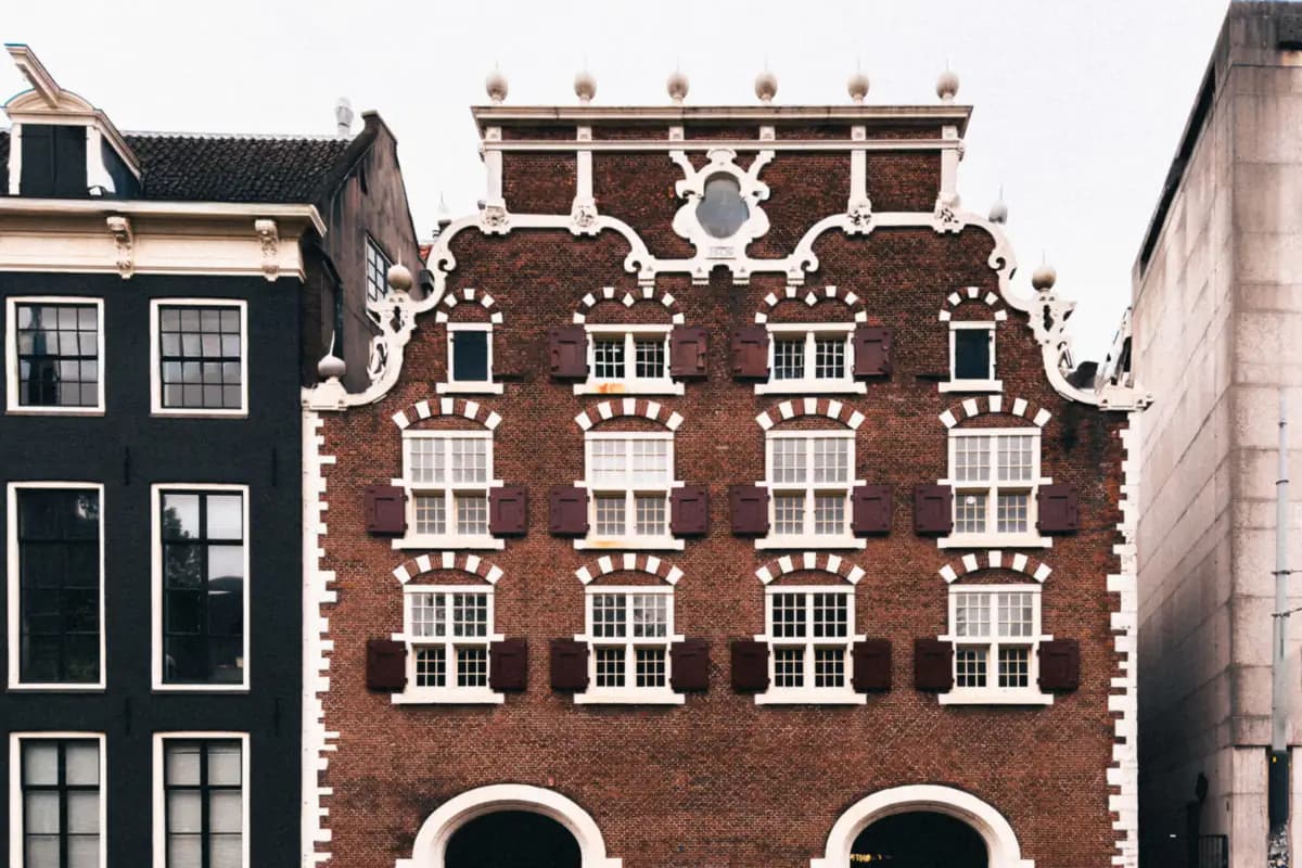 The brick front of an old building in Amsterdam with a white scrolled gable, red shutters and two arched doorways