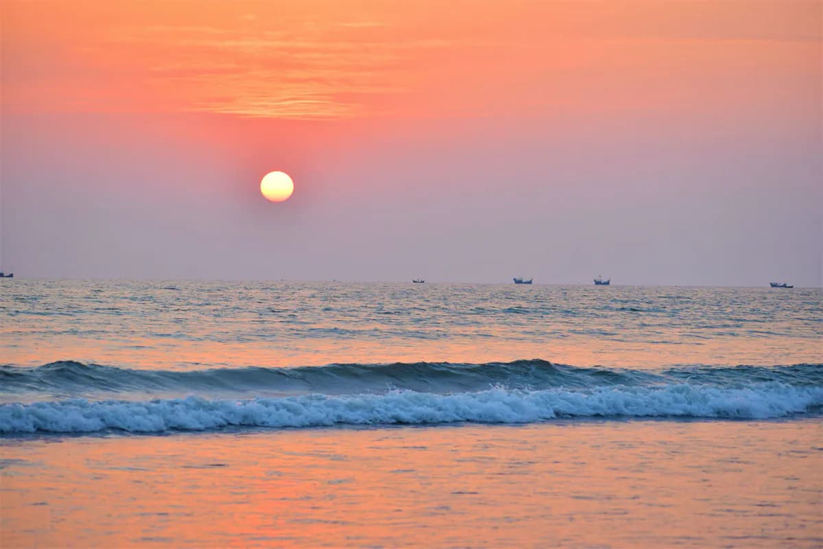 The sun setting over the Bay of Bengal, with waves breaking in the foreground and fishing boats on the horizon